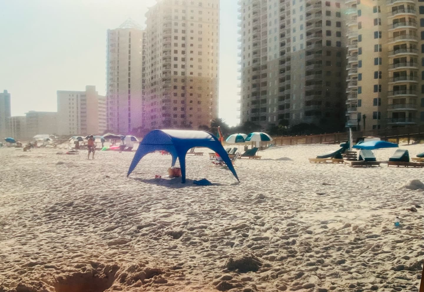 Archival photograph of an early Shady Baby prototype — a blue arched canopy pitched on the sand in front of beachfront high-rises