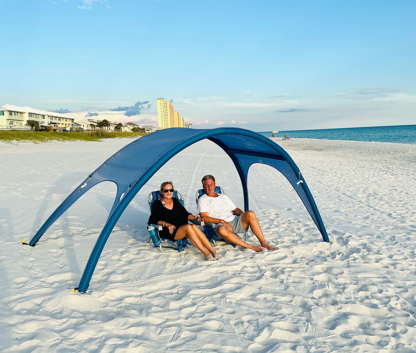 Shady Baby canopy staked on white sand, a couple seated in beach chairs in its shade with the ocean and shoreline behind
