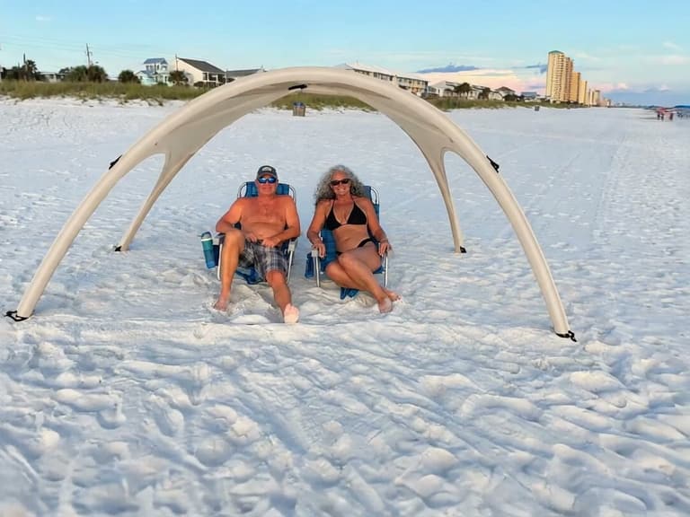 Shade Shack canopy in beige pitched on the beach at golden hour, two people seated in chairs beneath it